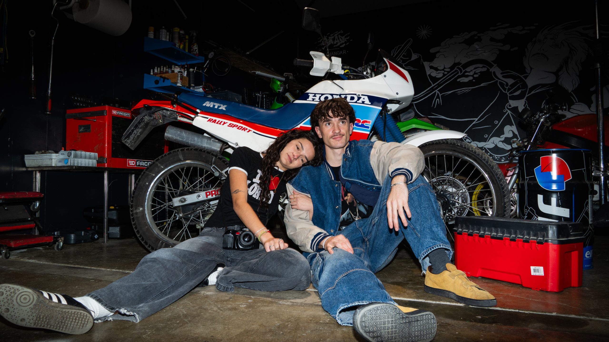 jeune couple habillé de façon vintage, qui pose devant une moto Honda dans un garage sombre
