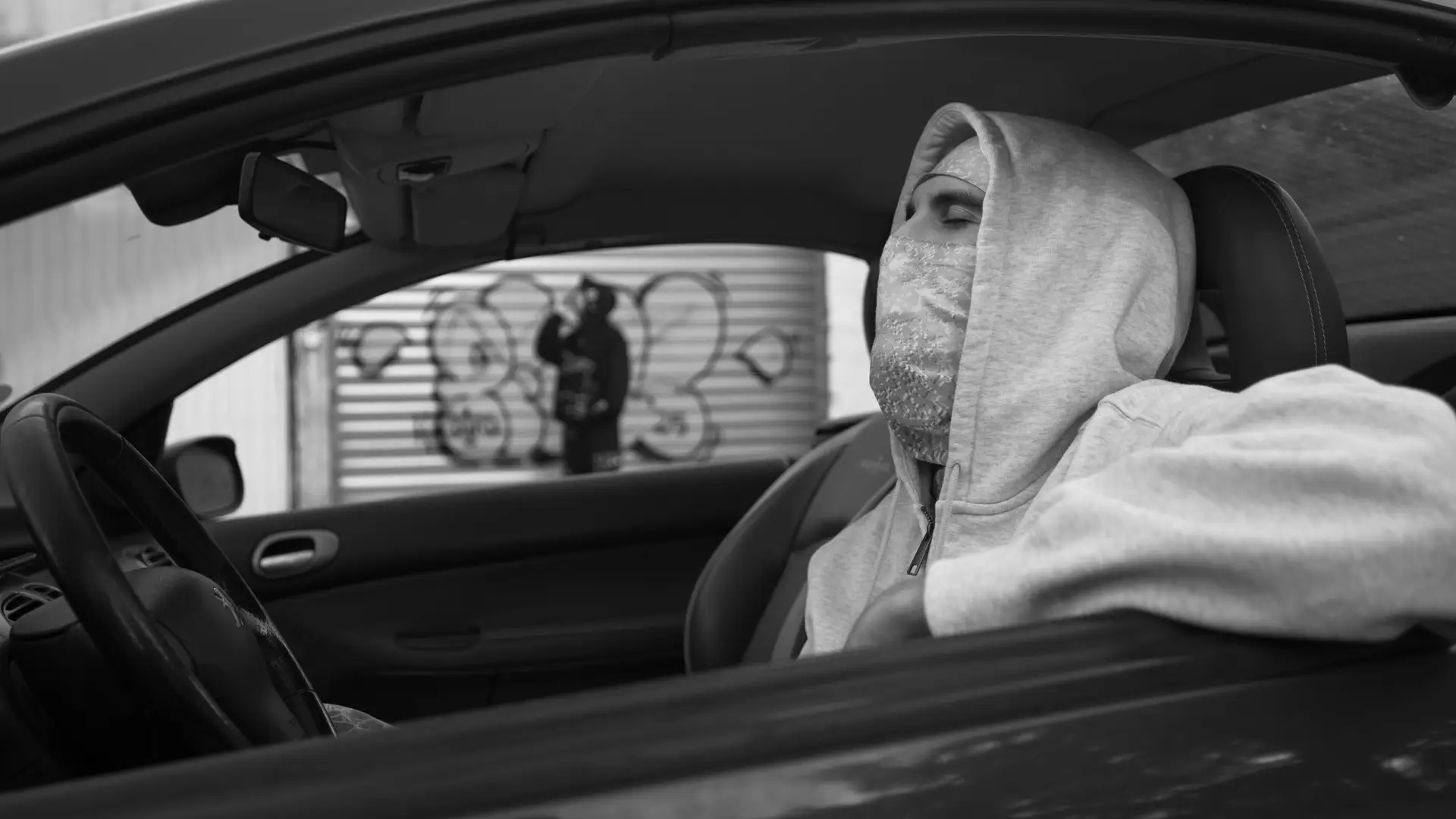 C'est une photo noir et blanc issue du clip Senzu de l'artiste Pivi avec Zelef. Pivi est cagoulé assis au volant de sa voiture, il a les yeux fermé. En arrière-plan Zelef est flou debout tout en noir devant un graffiti.