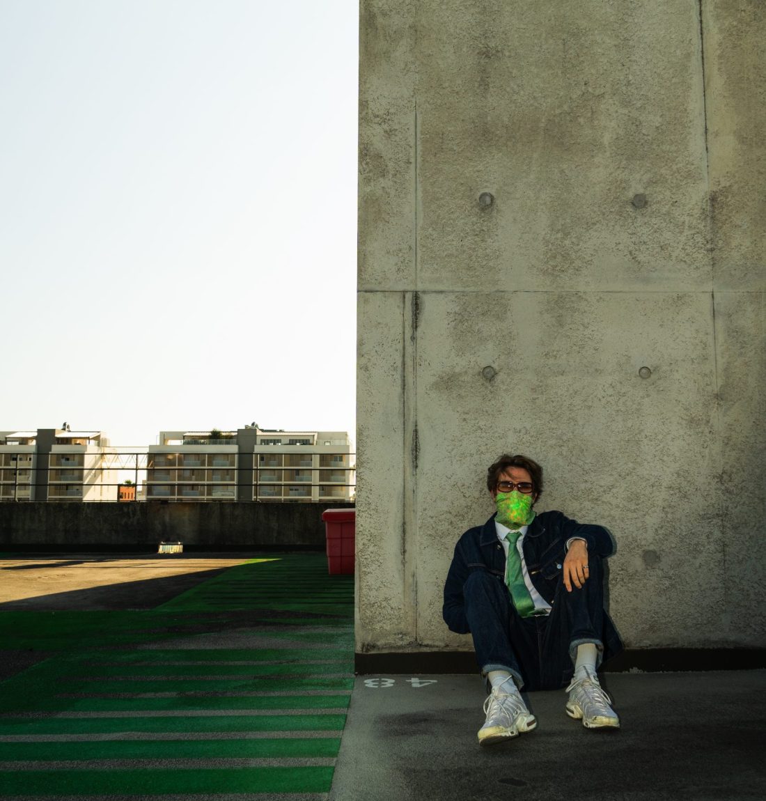 Un homme étant l'artiste Pivi est assis sur le sol contre un mur en béton brut, il porte des tn, un jean et une veste en jean brut, une chemise, une cravate verte, un masque au niveau du bas de son visage vert aussi et des lunettes de soleil. Il est assis en haut d'un immeuble, en voyant le haut de deux immeubles en arrière-plan. Il y a un passage piéton vert sur sa droite, ce qui laisse dire que la photo a été prise sur un parking.