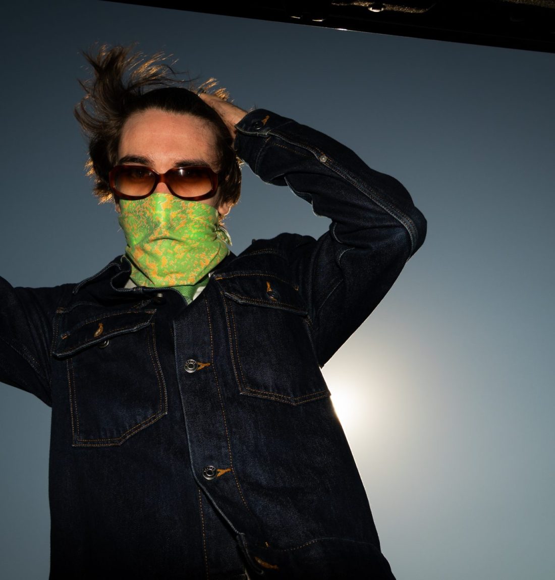 Un homme étant l'artiste Pivi se tient les cheveux en arrière, il porte des lunette de soleil et un masque vert et orange. Il est habillé avec une veste en jean, il tient avec sa main droite le haillon du coffre de sa voiture. Le soleil est dos à lui mais le flash de l'appareil photo l'éclaire ce qui donne un contraste très fort.
