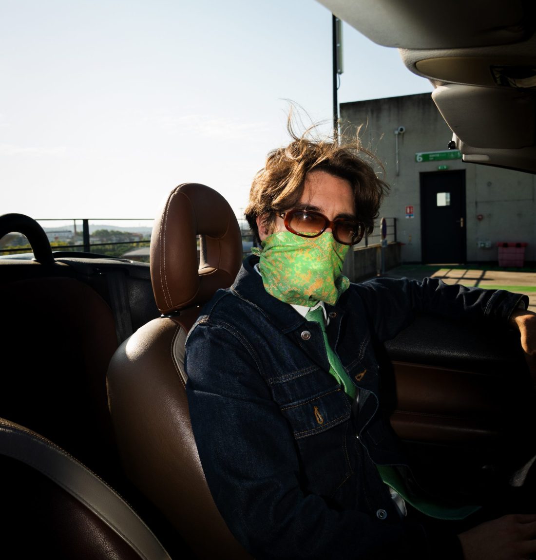Un homme étant l'artiste Pivi se tient assis sur le siège conducteur, il regarde la caméra avec ses lunettes de soleil. Il porte un masque pour caché le bas de son visage et une cravate verte avec une veste en jean.