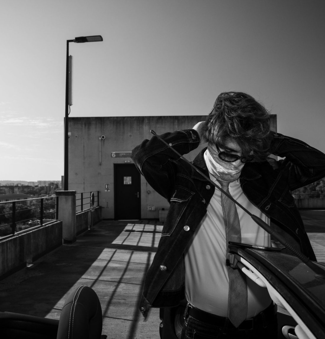 Une photo en noir et blanc de l'artiste pivi sur un parking il est devant sa voiture il accroche son bandana autour de son visage. On voit Toulouse en arrière plan de ce parking.