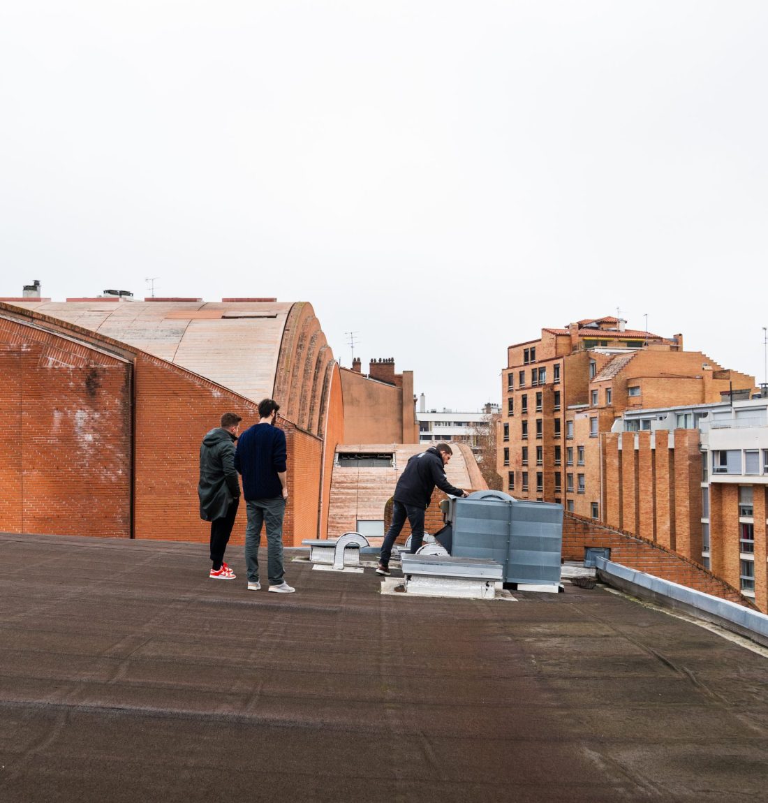 Ludovic de LP Ventil est sur le toit du centre de Toulouse, pour la création d'un devis pour une ventilation de cuisine d'un restaurant. Il regarde la ventilation et les clients sont à coté de lui. Il fait jour sur la photo, les briques rouges toulousaines ressorte fortement.