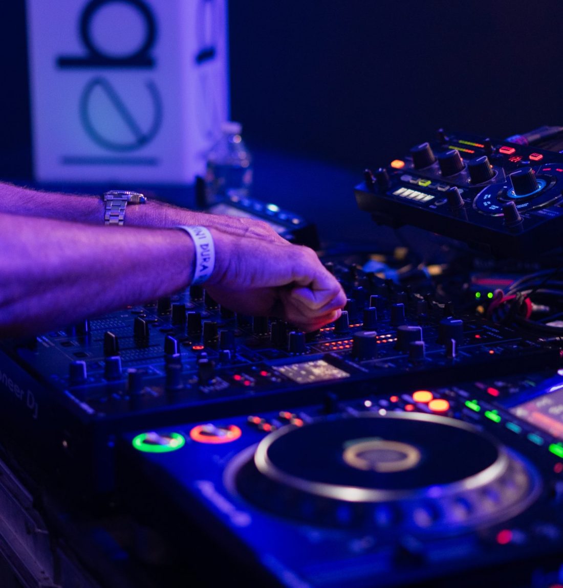 Une table de mixage est utilisé pour un concert. Les couleurs sont dans des tons violets et bleus, un homme est entrain de mixer avec ses deux mains dessus.