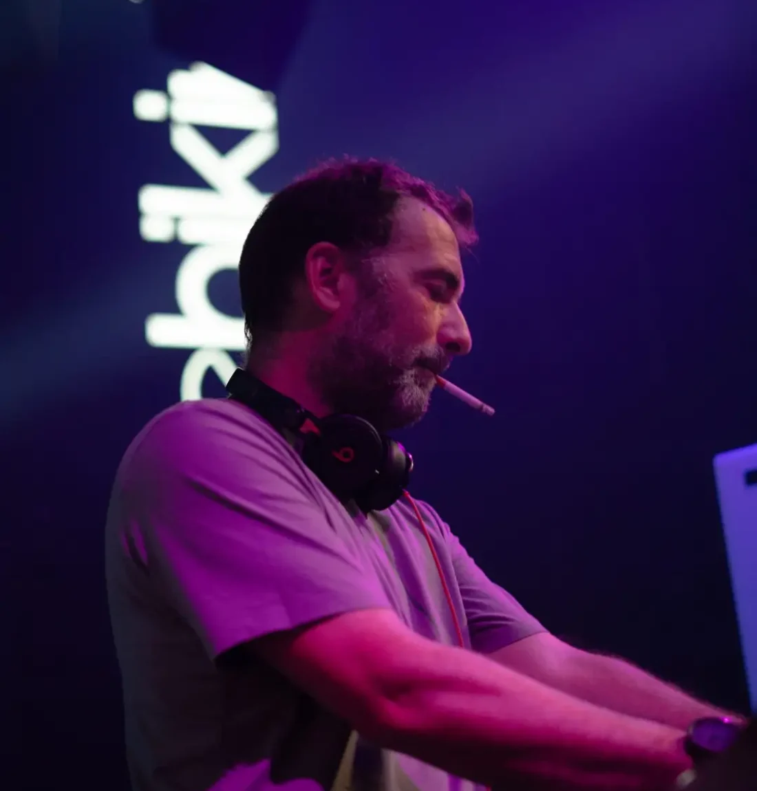 Un des deux membres du groupe The Deep Brothers est entrain de mixer on voit en arrière plan le logo de la salle de concert toulousaine le Bikini. Il fume et a un casque beats autour du cou.
