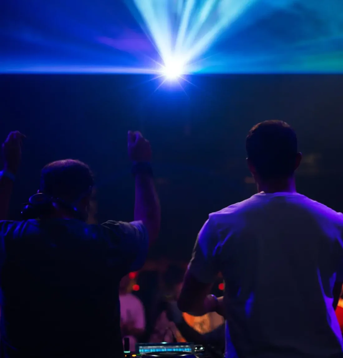 Un grand halo bleu venant d'une lumière prend toute la partie haute de l'image. On voit les contours des deux dj de dos entrain de mixer et de s'ambiancé et le public derrière entrain de danser.