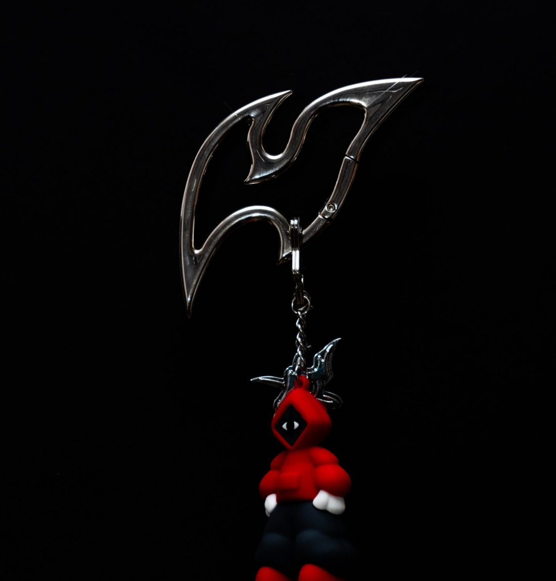 Sur un fond noir uni, nous voyons en détail le produit de la marque citizen. C'est un mousqueton avec une forme peu commune et assez pointu. Accroché il y a un porte clef représentant un bonhomme portant un sweat rouge, c'est la mascotte de la marque.