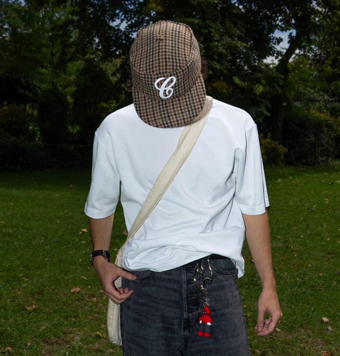 Un homme baissant la tête et se faisant cacher par sa casquette à damiers. Il porte un tee shirt blanc, un jean noir, une sacoche et un porte clef de la marque citizen étant constitué d'un mousqueton dans la forme du logo de la marque et un bonhomme rouge étant la mascotte de la marque. Il est debout dans un parc avec des arbres en arrière-plan. Arrère-plan sombre par rapport au sujet par le flash de l'appareil photo.