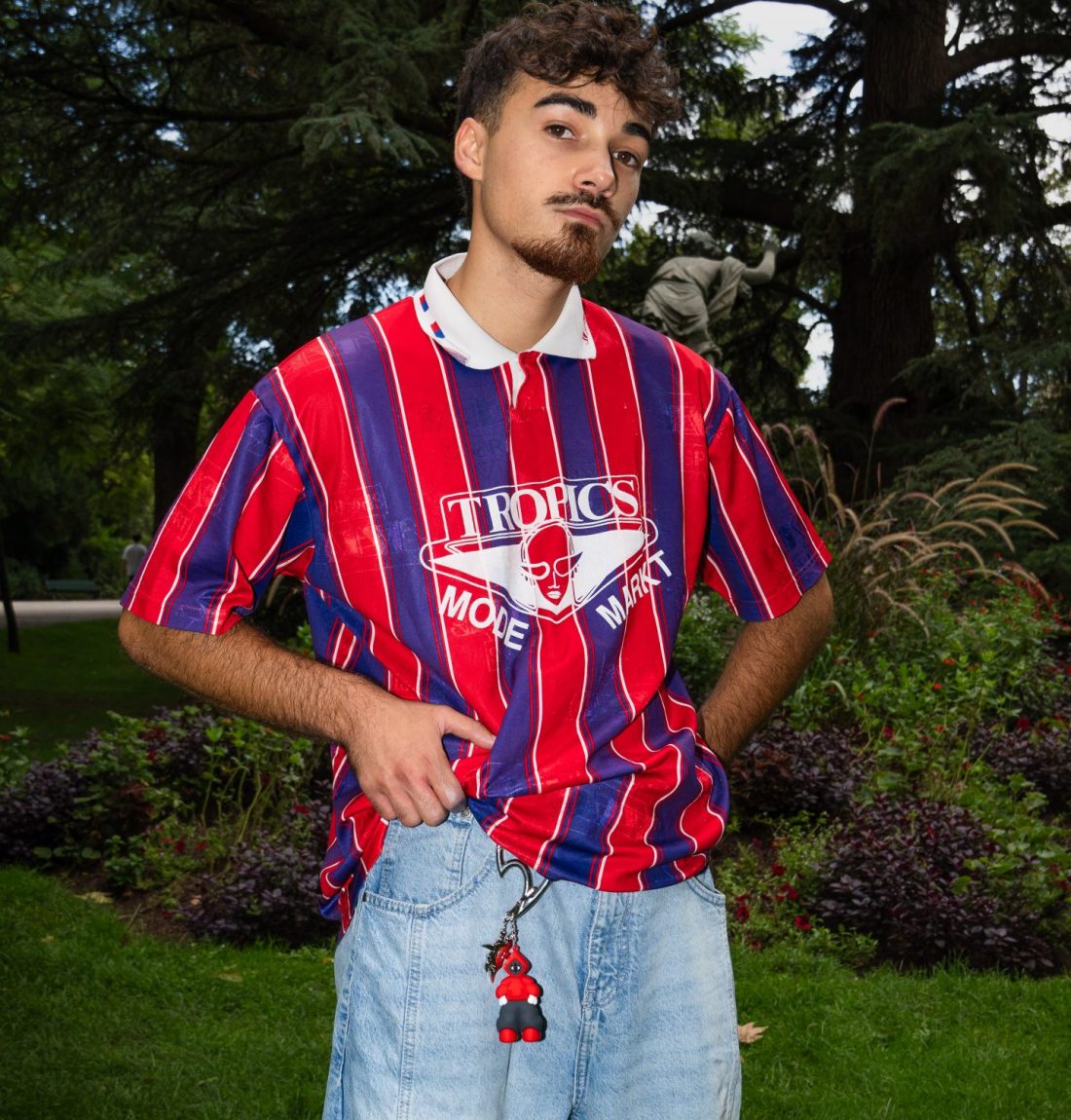 Un mannequin masculin regarde la caméra il relève légèrement avec sa main droite son polo à rayure rouge et violette pour laisser apercevoir un mousqueton de la marque citizen ayant accroché un porte-clef représentant la mascotte de la marque. Il est debout dans un parc devant des arbres l'arrière-plan est très sombre pour que le mannequin ressorte avec le flash.