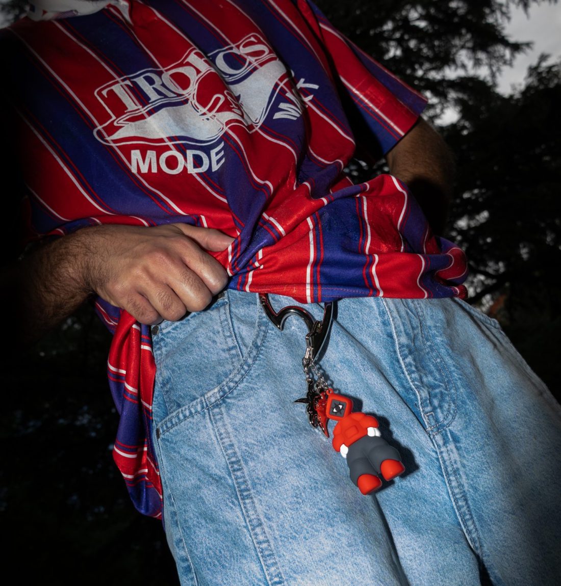 Un homme dans l'obscurité soulève son polo et en premier plan on voit accroché à son jean bleu, un mousqueton de la marque citizen et sa mascotte accroché en porte-clef dessus.