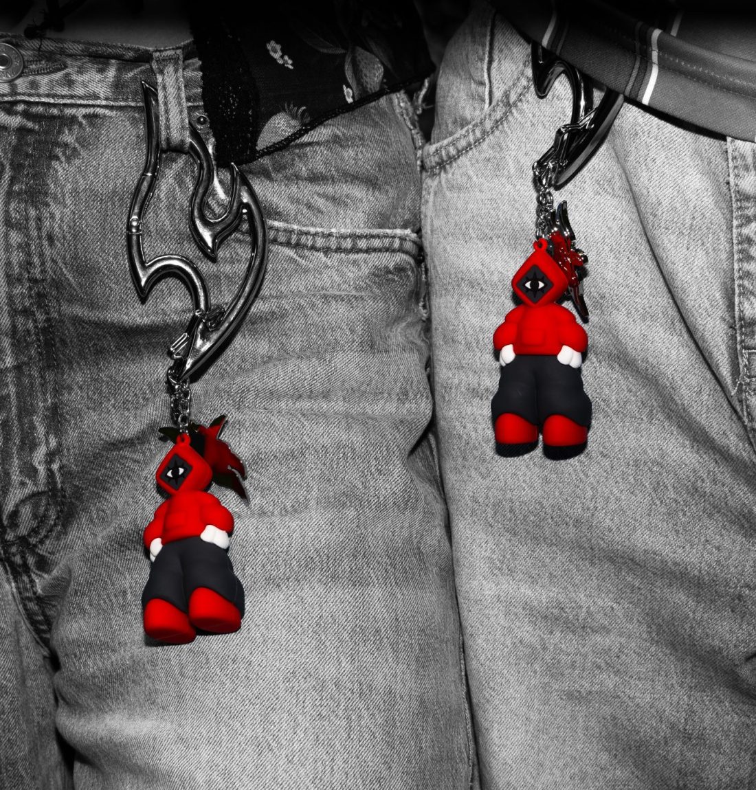 C'est une photo focalisé sur le haut de deux jeans. La photo est en noir et blanc excepté le détail d'un porte-clef accroché au jean à l'aide d'un mousqueton. Cet ensemble est un accesoire de la marque citizen. Le détail en couleur est la mascotte portant un sweat rouge.