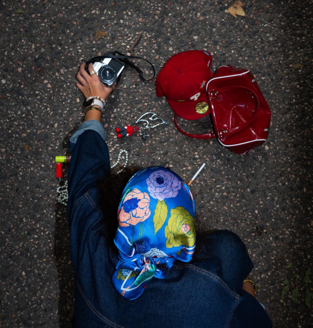 Nous voyons sur un sol bétonné un sac à main rouge renversé avec une casquette rouge, un briquet, des bijoux un appareil photo, une cigarette et un porte-clef avec un mousqueton de la marque citizen. Une personne tout en bleu est accroupi et attrape l'appareil photo.