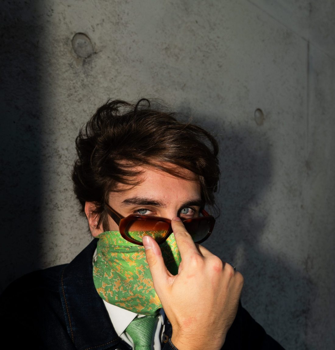 L'artiste Pivi regarde la caméra avec ses yeux bleus et tient ses lunettes de soleil sur son nez il est contre un mur de béton brut et le flash donne un aspect artistique à la photographie.