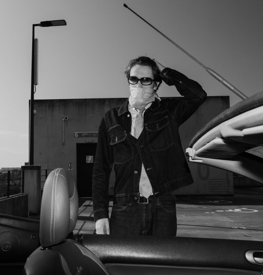 Une voiture décapotable en tout premier plan, l'artiste Pivi est derrière cette voiture en ensemble jean. C'est une photo noir et blanc.