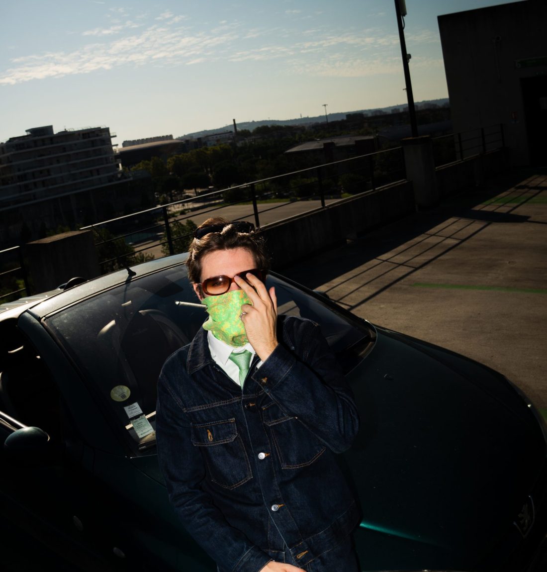 Une photo grand-angle, l'artiste pivi se cache la moitié du visage avec sa main et porte des lunettes de soleil, un bandana autour du bas de son visage, un ensemble en jean et une cravate verte. Il est assis sur l'avant de sa voiture. Il fait jour mais la lumière est très sopbre pour faire ressortir le premier plan éclairé par le flash. Il est sur le haut d'un parking à Toulouse on distingue le zénith en arrière-plan.