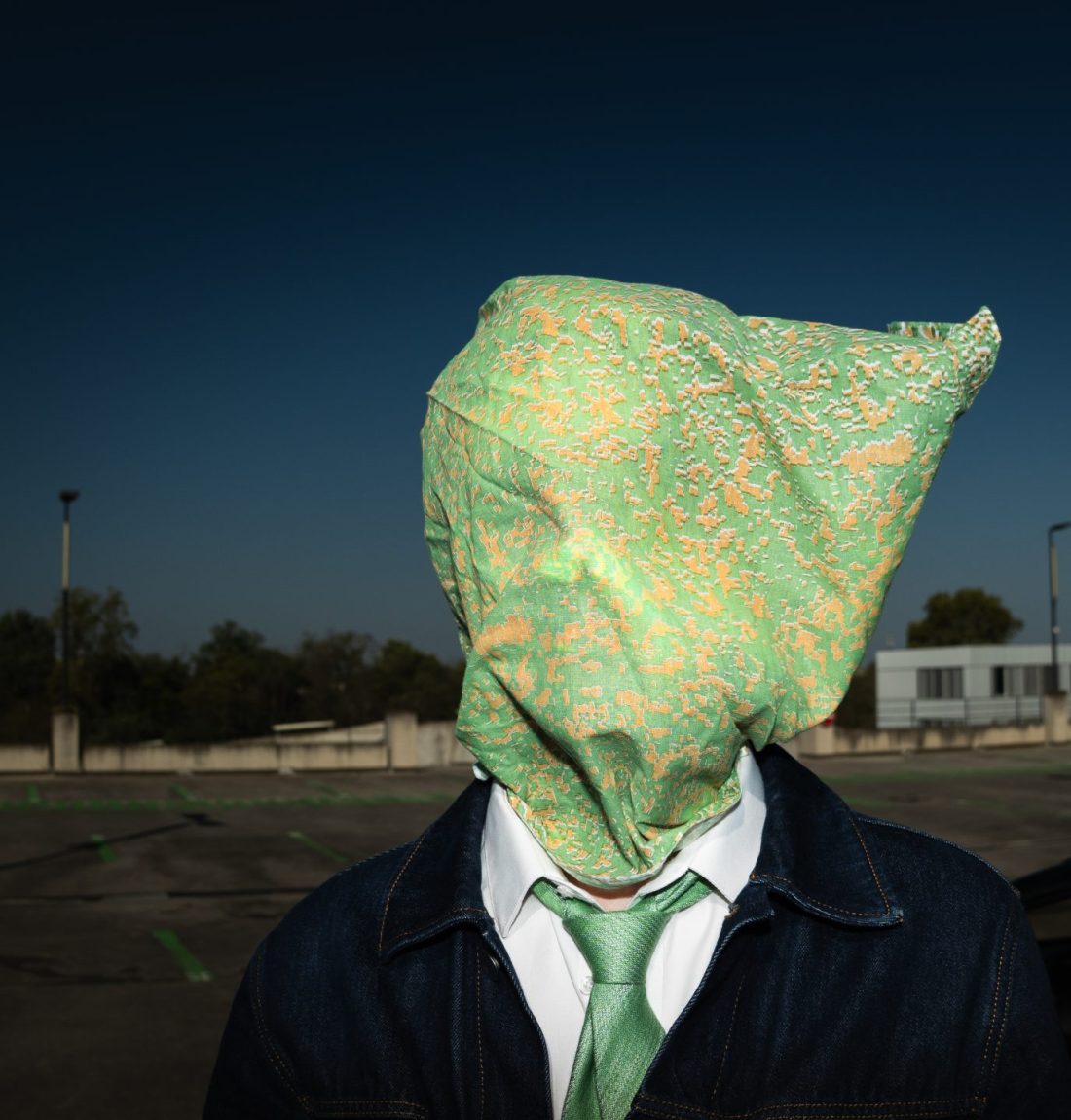 Un homme sur un parking a le visage totalement caché par un bandana vert et orange. Il porte une cravate verte et une veste en jean. Il est sur un parking vide. L'arrière plan est très sombre mais le sujet est très bien éclairé.