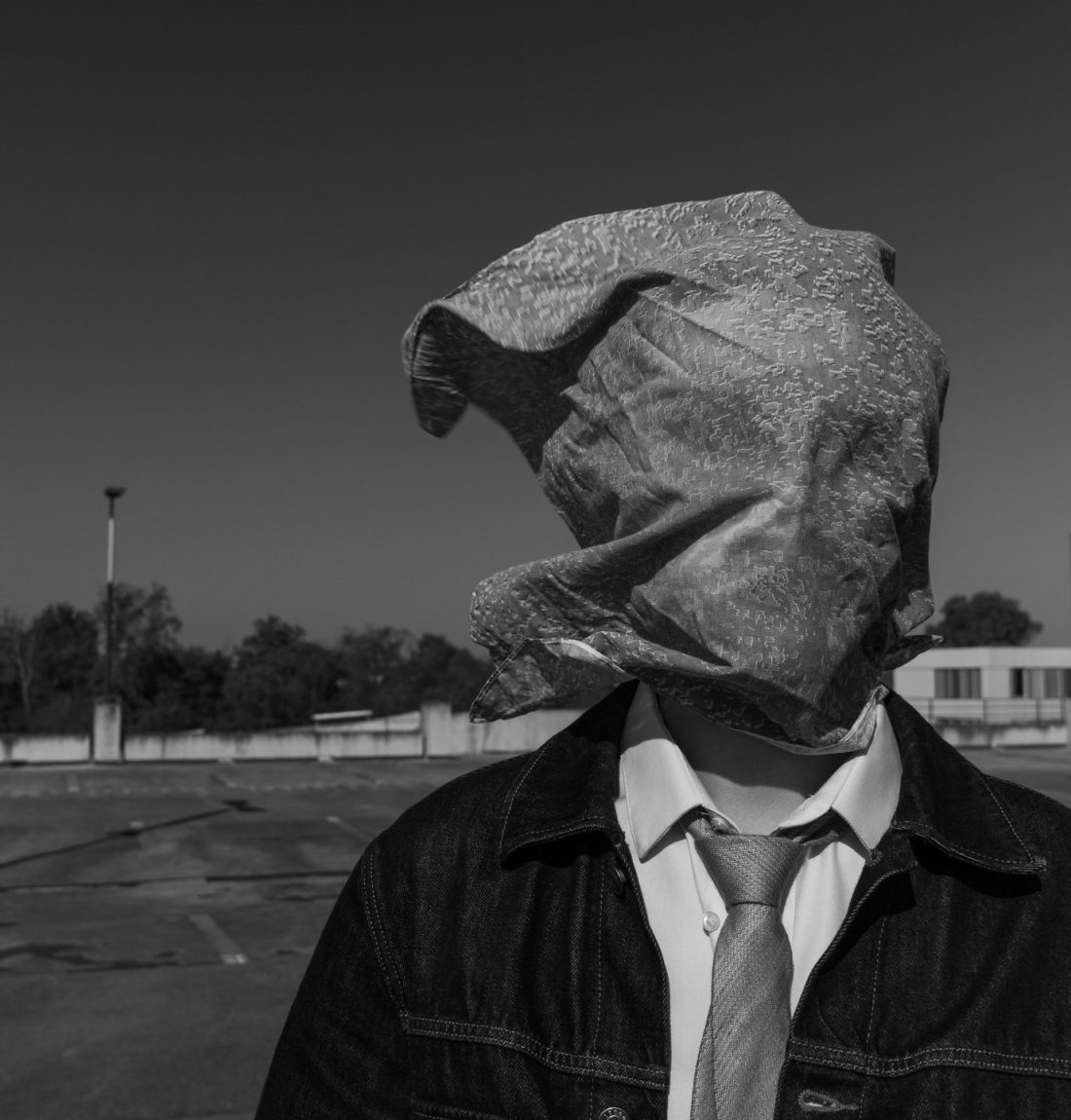 Un homme sur un parking a le visage totalement caché par un bandana vert et orange. Il porte une cravate verte et une veste en jean. Il est sur un parking vide. L'arrière plan est très sombre mais le sujet est très bien éclairé. C'est une photo en noir et blanc ce qui dé-contraste légèrement la photo.