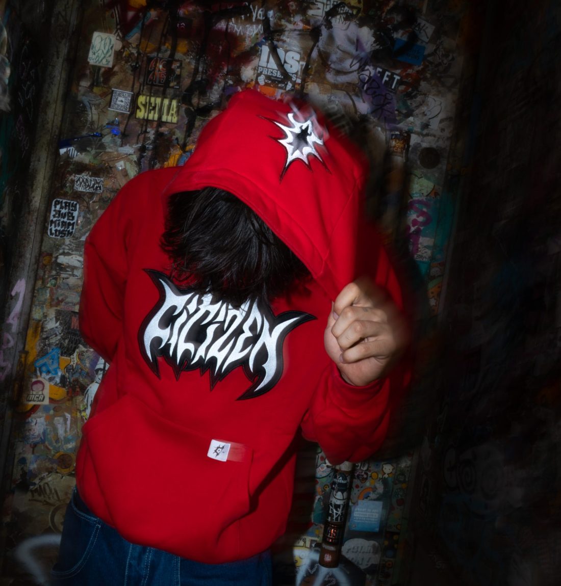 Un homme tire sa capuche vers le bas, ce qui montre l'ensemble des détails de broderie de son pull de la marque citizen. La photos est très contrasté avec un léger effet de flou.