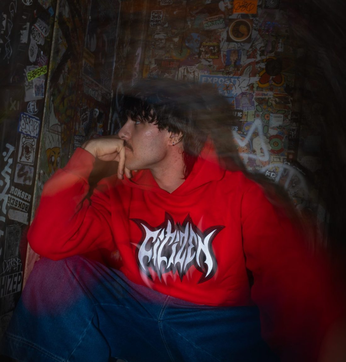 Un mec est assis sur une marche il regarde sur sa droite en étant posé sur sa main, il est dans l'obscurité et est éclairé par le flash de la photo, un fort flou artistique domine la photo. Il porte un sweat rouge de la marque citizen