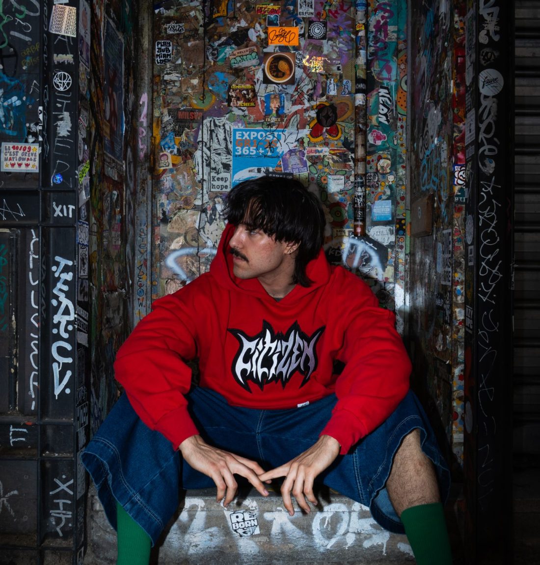 jeune homme devant porte graphée avec pull rouge citizen il prend la pose et le pull rouge ressort du fond assez sombre