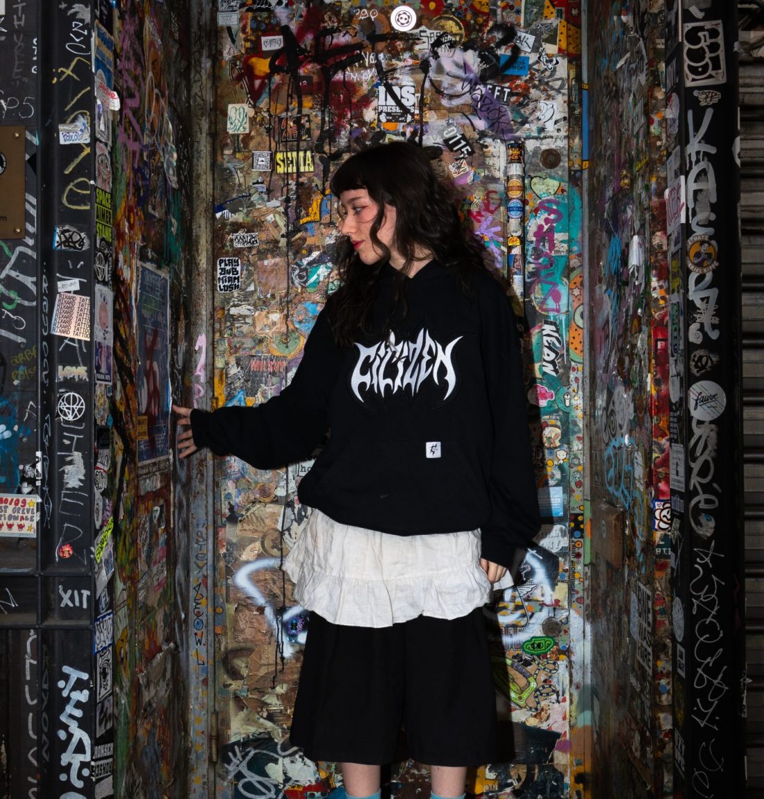 Une fille qui porte un sweat noir de la marque citizen, deux robes est devant une porte rempli de graffiti et d'autocollants, elle touche le mur avec sa main droite.