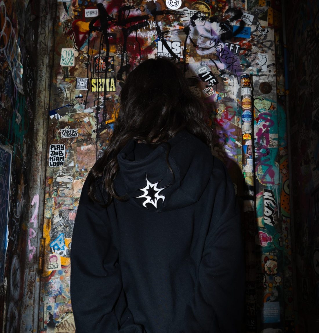 Une fille de dos devant un coin graffer de partout, elle porte un pull noir de la marque citizen, on voit le détail de broderie sur la capuche. Elle se tient les mains.