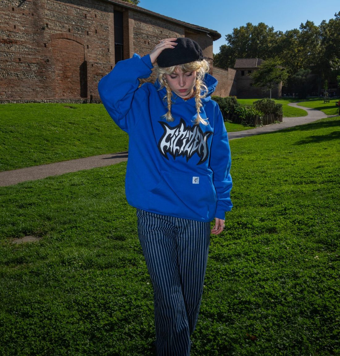 Une femme blonde se tient debout les jambes croisés elle regarde vers le bas avec sa main droite sur le chapeau noir qu'elle porte sur sa tête. Elle porte aussi un sweat à capuche bleu de la marque citizen. Les couleurs sont très contrastés mais elle ressort de l'image grâce au flash de la photo.