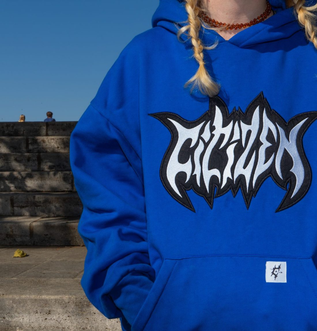 Le pull bleu de la marque citizen est en premier plan et prends la moitié de l'image. On voit très bien les détails de broderie et du logo sur l'avant du pull. Deux nattes blondes de la mannequin tombe sur le pull.