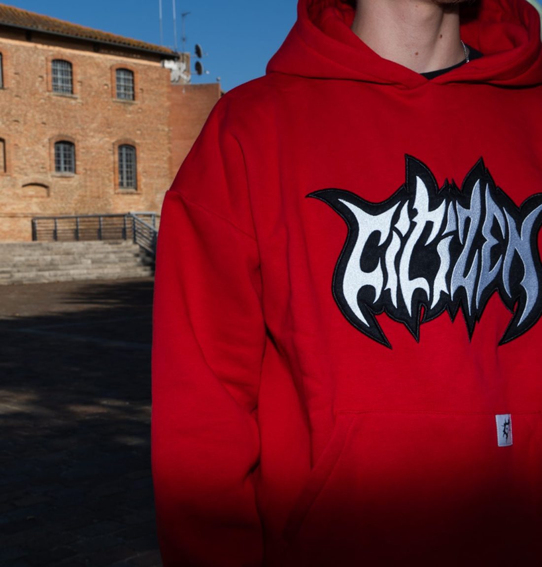 Le pull rouge de la marque Citizen prend la moitié du cadre. Le sweat ressort énormément grâce au flash de la photographie et ressort des ombres de l'arrière-plan. Un batiment en brique rouge est présent sur l'arrière plan et comble le vide sur la partie gauche de l'image.