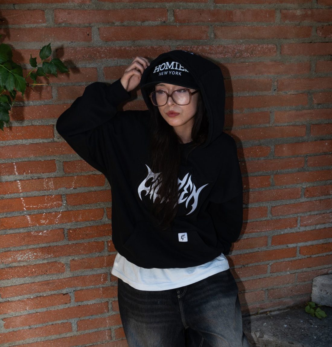 Une jeune femme à lunette est adossé contre un mur de brique au pied de marche. Elle porte un sweat noir Citizen, elle tient sa capuche par la main droite et regarde sur sa droite. Elle porte aussi une casquette, un tee-shirt blanc dépasse de son pull et un jean large noir. La photo est éclairé grâce à un flash qui donne un certain effet artistique.