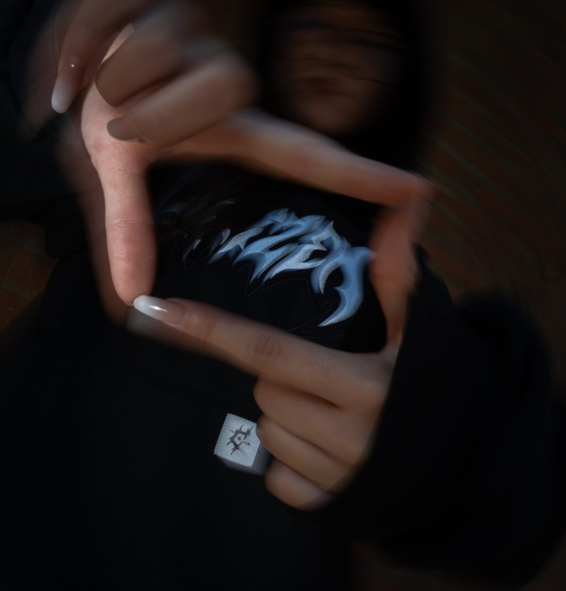 Une fille à lunette tend ses deux bras vers la caméra est forme un rectangle avec ses doigts. À l'intérieur du cadre qu'elle crée on voit la broderie du logo du pull noir citizen. La photo est très sombre avec un flou de mouvement ce qui laisse la place aux détails blancs de la broderie sur le pull.
