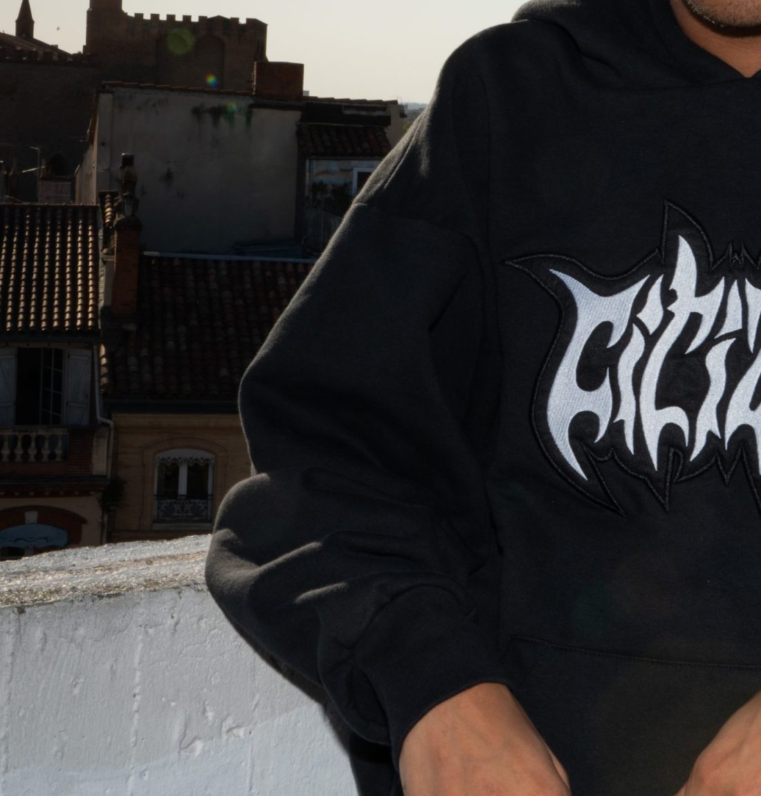 Le pull noir de la marque Citizen prend la moitié de l'image, sur le restant de l'image on voit que le mannequin portant le pull est adossé à un muret et derrière ce muret on voit le haut de bâtiment en briques avec des tuiles rouges. L'arrière-plan est très sombre dû au flash de l'appareil photo qui sert à faire ressortir le pull que le mannequin porte.