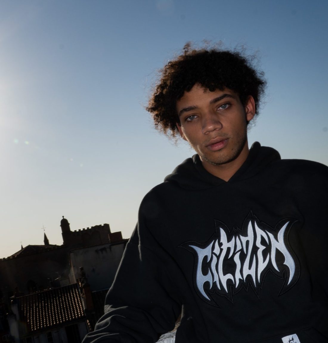 Un homme regarde la caméra, il est à contre-jour mais le flash de la photo permet de très bien le voir. Il porte un sweat noir de la marque citizen, on voit le détail de la broderie du sweat, où il y a écrit citizen sur le buste. Le mannequin est aussi adossé sur un muret.
