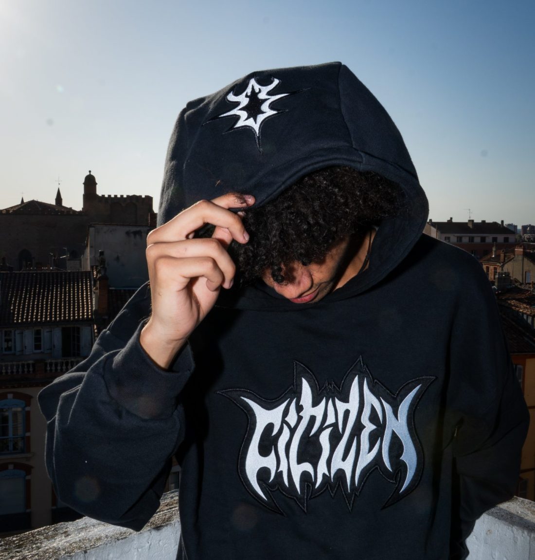 Un jeune homme tient avec sa main droite la capuche de son pull. Ce pull est un pull noir de la marque Citizen, il y a beaucoup de détail de broderies, dont un grand texte écrit citizen sur le buste et sur le haut de la capuche un signe noir et blanc. Il est debout posé en haut d'un immeuble à contre-jour, le flash de l'appareil permet de le détacher de l'arrière plan très sombre.