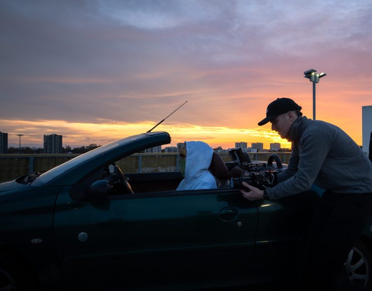 Le rappeur est garé il est assis dans sa voiture décapotable verte, il pose pour le cadreur qui est entrain de filmer dans le rétro de la voiture, on peut apprécier le beau coucher de soleil en arrière plan