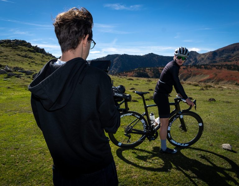 Photo d’un cadreur tenant une caméra Sony montée sur un stabilisateur DJI Ronin, dans un décor naturel avec des montagnes aux couleurs rouges et vertes, Le cadreur filme un cycliste professionnel, on voit son vélo en carbone. Cette image montre l’envers d’un tournage avec un cycliste professionnel.
