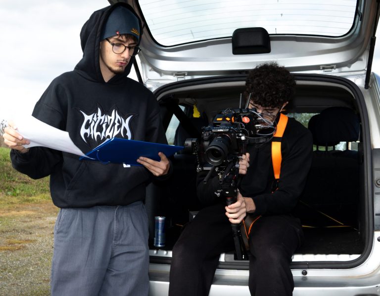 Deux personnes près du coffre: une personne assise dans le coffre avec stabilisateur et caméra + écran; autre en dehors portant sweat noir et bonnet bleu, dossier bleu; ambiance tournage, canette énergétique; arrière-plan verdure et ciel nuageux.