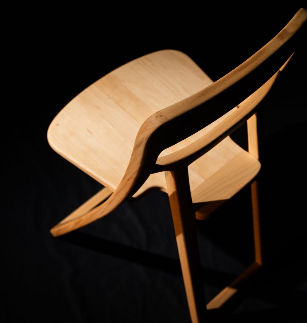 Chaise en bois clair très travaillée, elle est mise en valeur par l'éclairage, la photo est prise en plongée sur fond noir