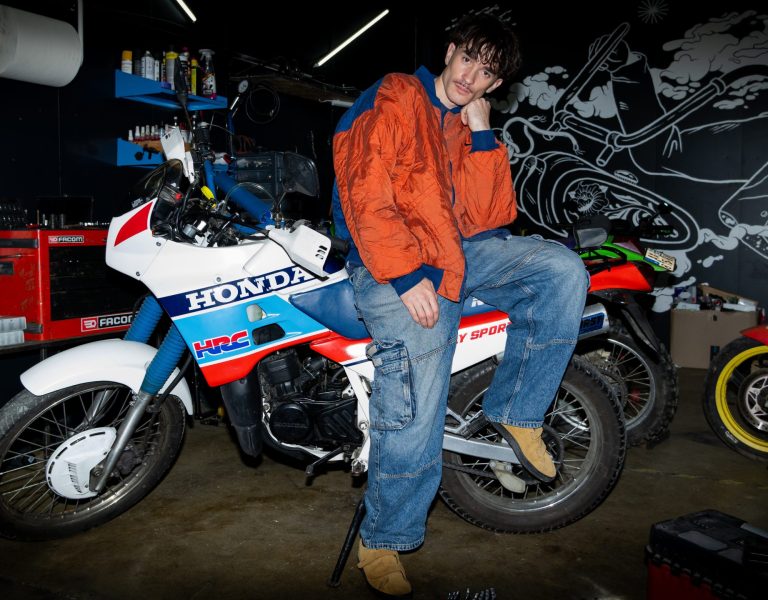 Atelier mécanique: homme en veste orange sur une moto Honda; outils FACOM et étagères; mur avec fresque motarde; ambiance d’atelier.