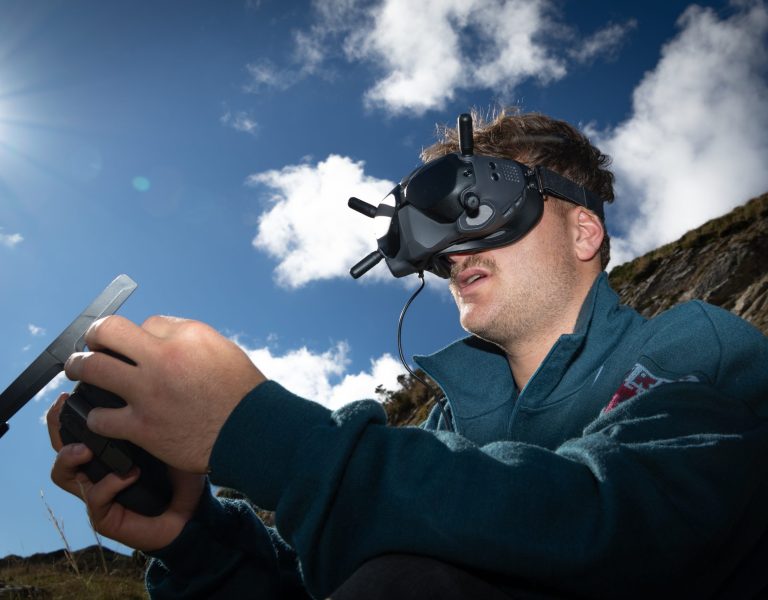 Photo d’un pilote de drone tenant une télécommande et portant des lunettes fpv dji google, on voit un joli ciel bleu percé d’un soleil puissant, et la montagne en arrière plan, le droniste est concentré cela montre la maitrise de ce type de prises de vue drone dans un milieu hostile qui est la montagne.
